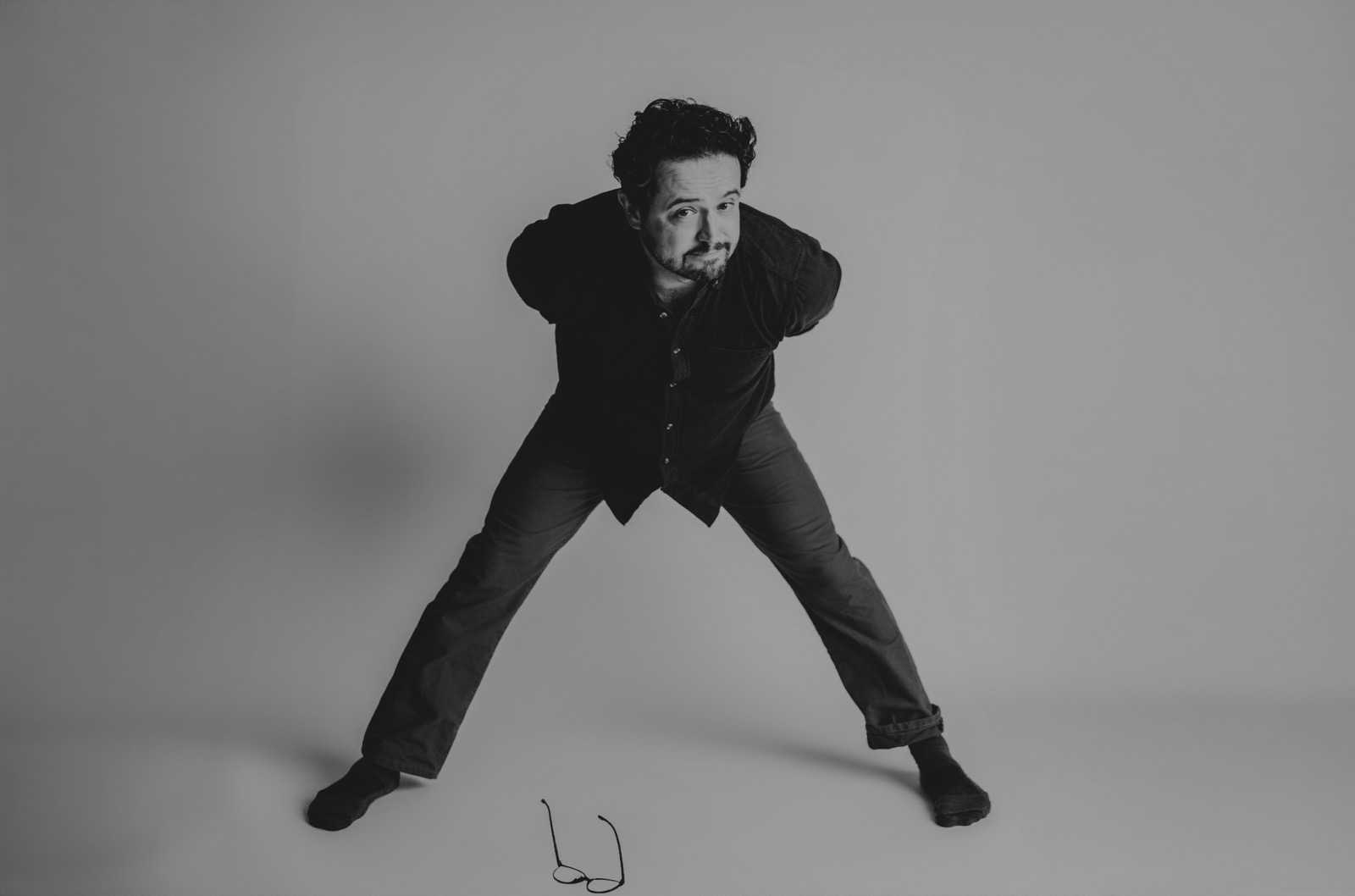 Black and white frame of Aaron braced in a wide stance with both hands behind the head, leaning in toward the camera in socks, glasses lying on the studio floor in front