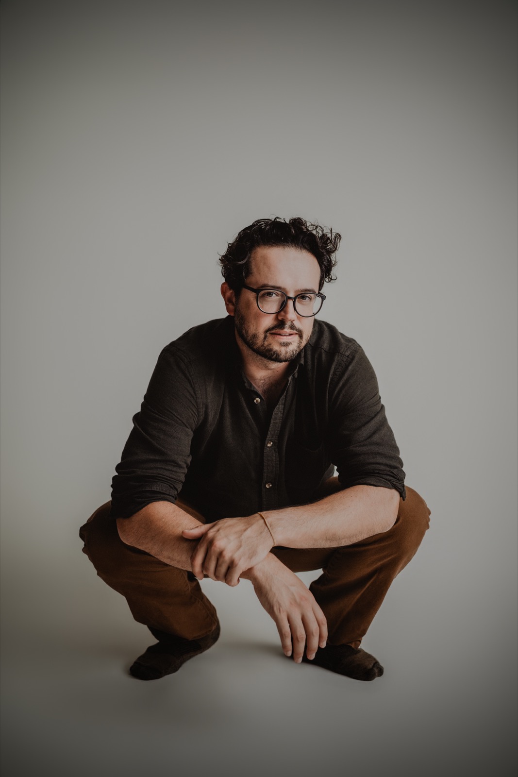 Aaron May crouched low with forearms resting on knees, glasses on, looking straight into the camera against a plain studio backdrop