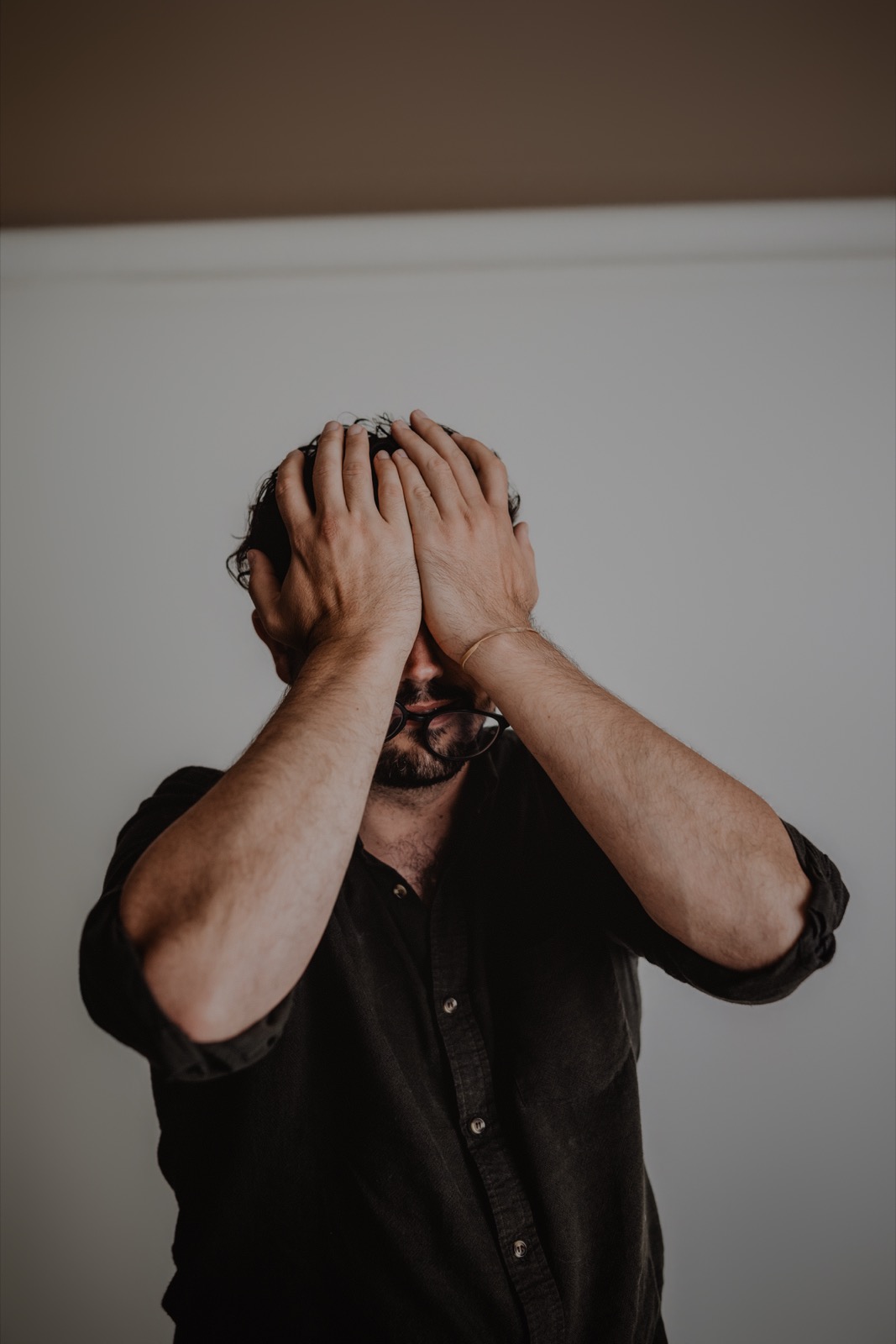 Aaron standing against a pale backdrop with both hands covering the face, only glasses and hair visible above the fingers