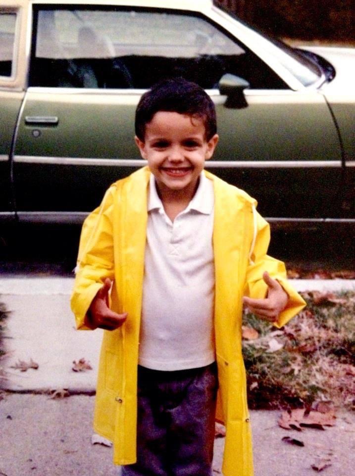 Aaron as a small boy in a bright yellow raincoat several sizes too big, grinning at the camera on a dry suburban sidewalk in front of a green station wagon
