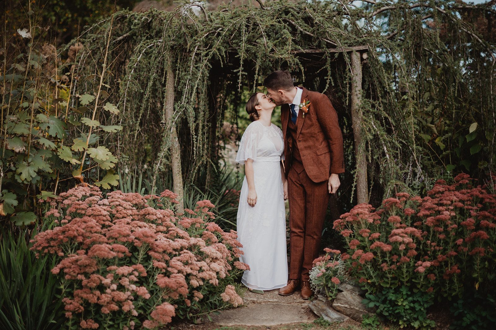 A couple kissing beneath a weeping larch arch in a garden, banks of dusty pink sedum on both sides of the path