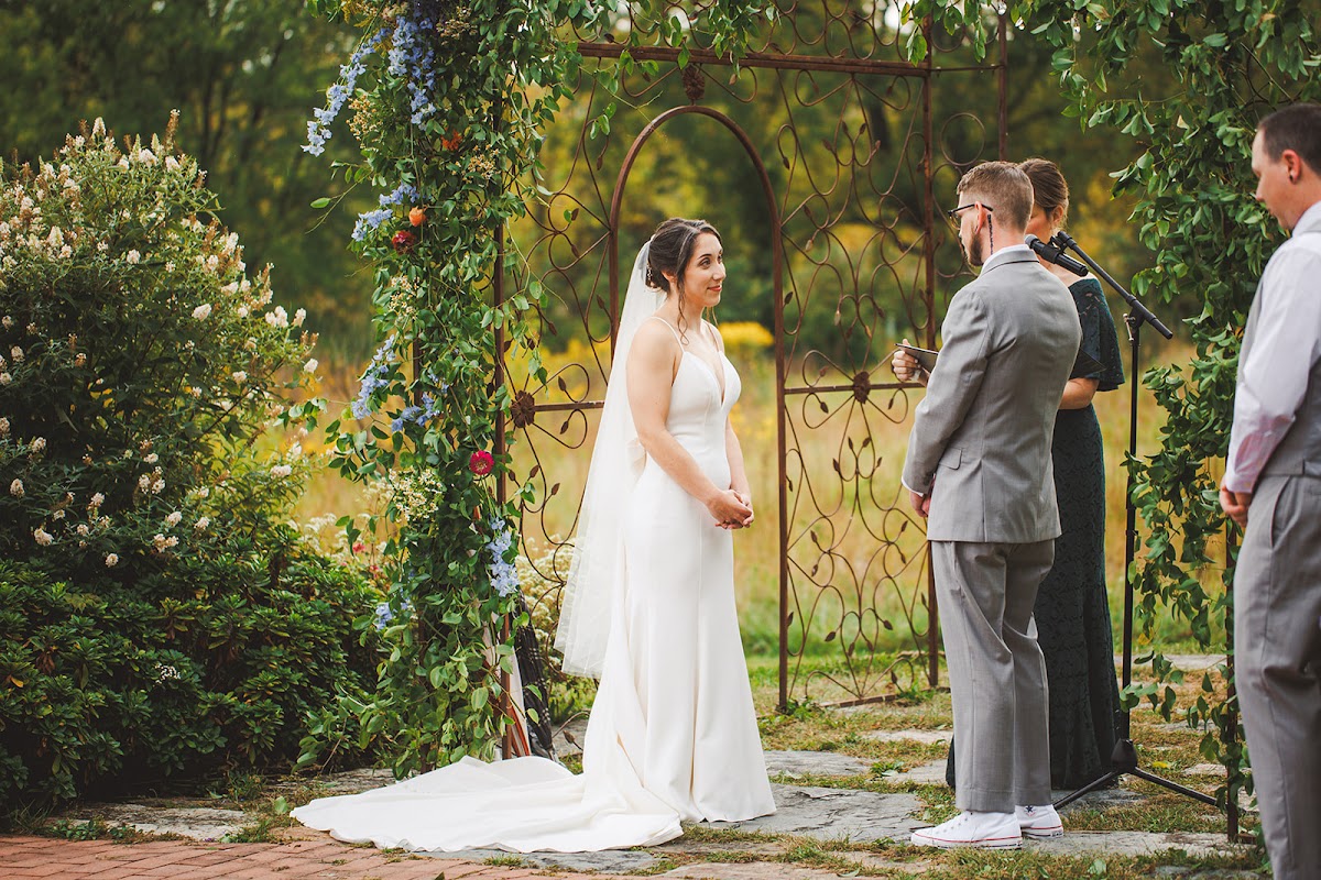 Amie in her veil standing at the vine-wrapped iron arbor during the ceremony as the officiant reads, golden meadow beyond