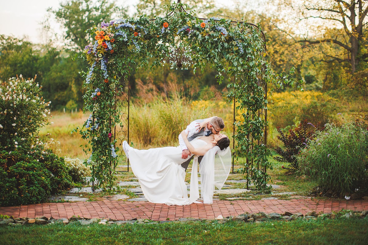 Cody dipping Amie for a kiss beneath the flower-topped metal arbor at Zigbone Farm Retreat, a small chandelier hanging above them and golden grasses behind
