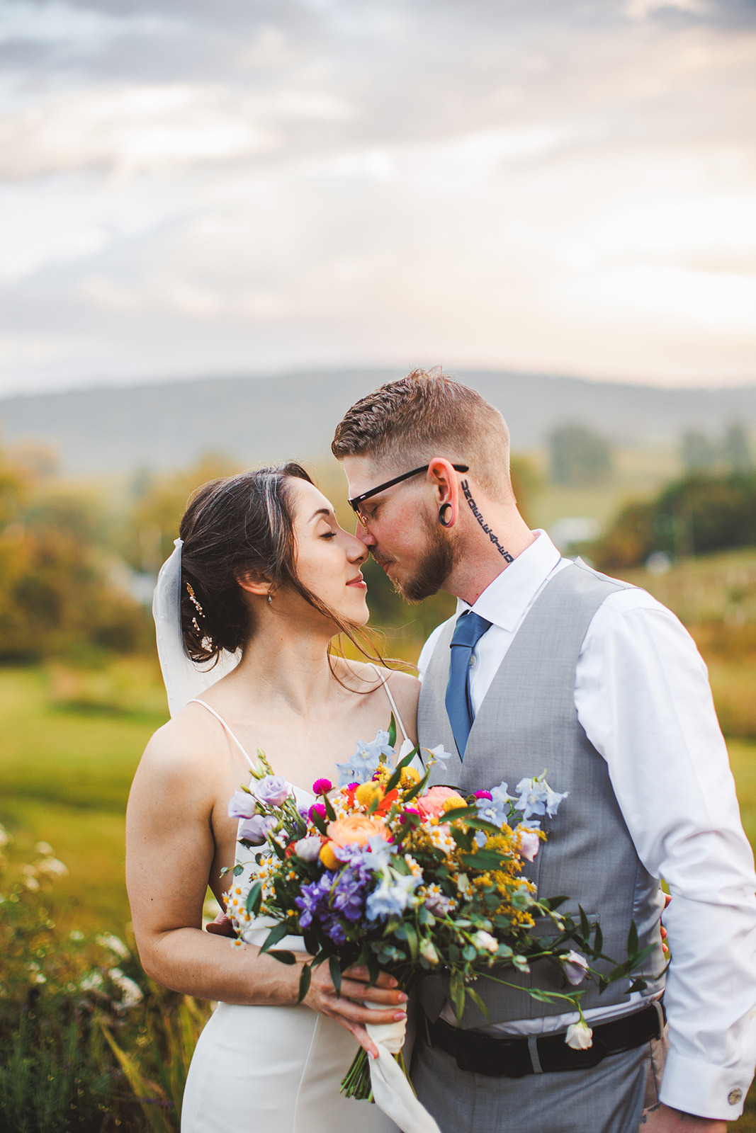 Amie and Cody nose to nose in the hillside meadow, her wildflower bouquet between them, rolling farmland and a dramatic evening sky behind
