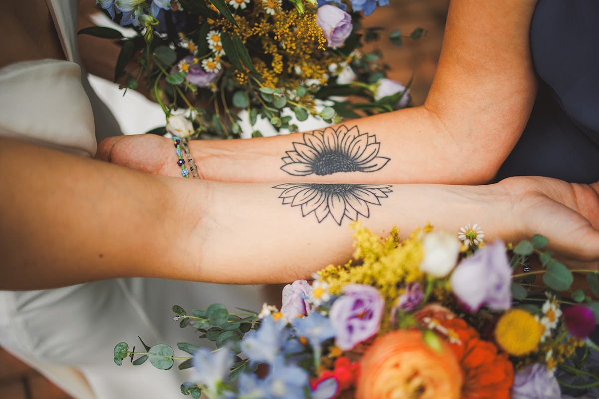 Amie's and Cody's forearms side by side showing matching sunflower tattoos, wildflower bouquets filling the frame around them