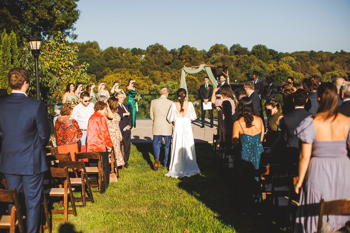 Andrea walking down the grass aisle between standing guests toward the draped arch, the wooded valley beyond the terrace