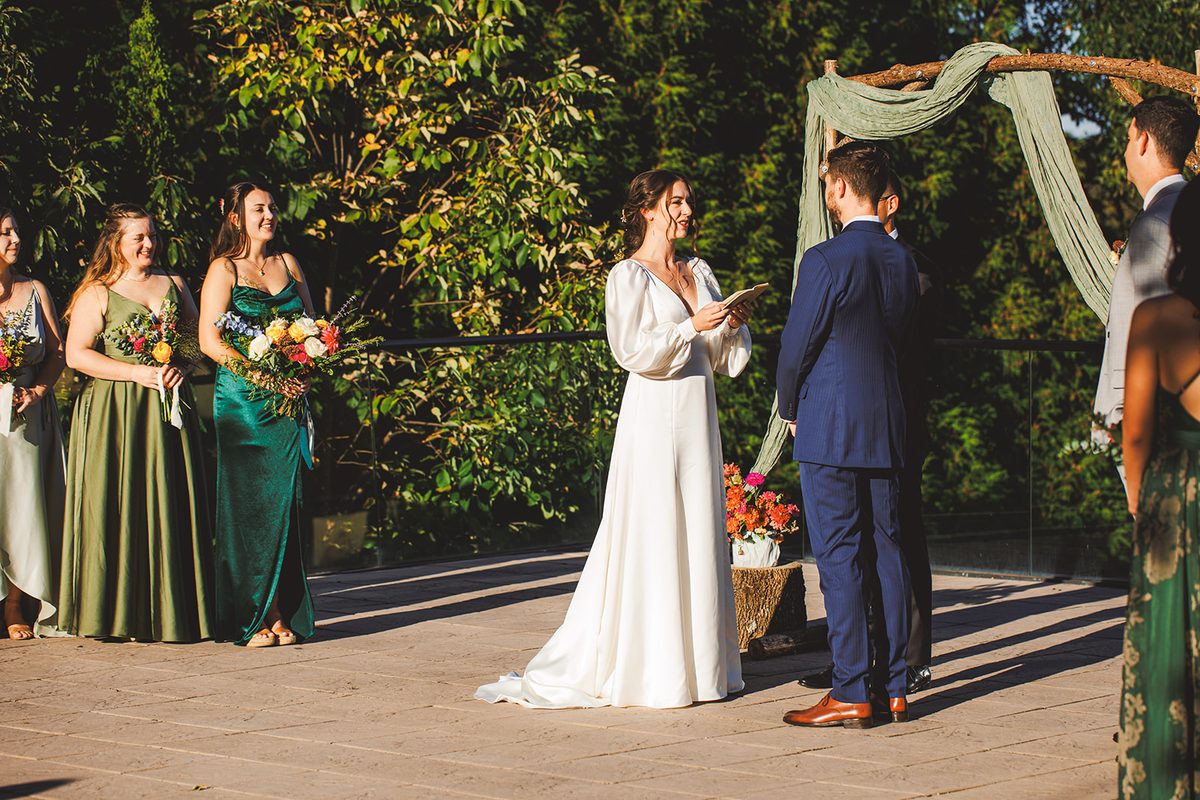 Andrea smiling as she reads her vows, her bridal party in shades of green watching from beside the arch