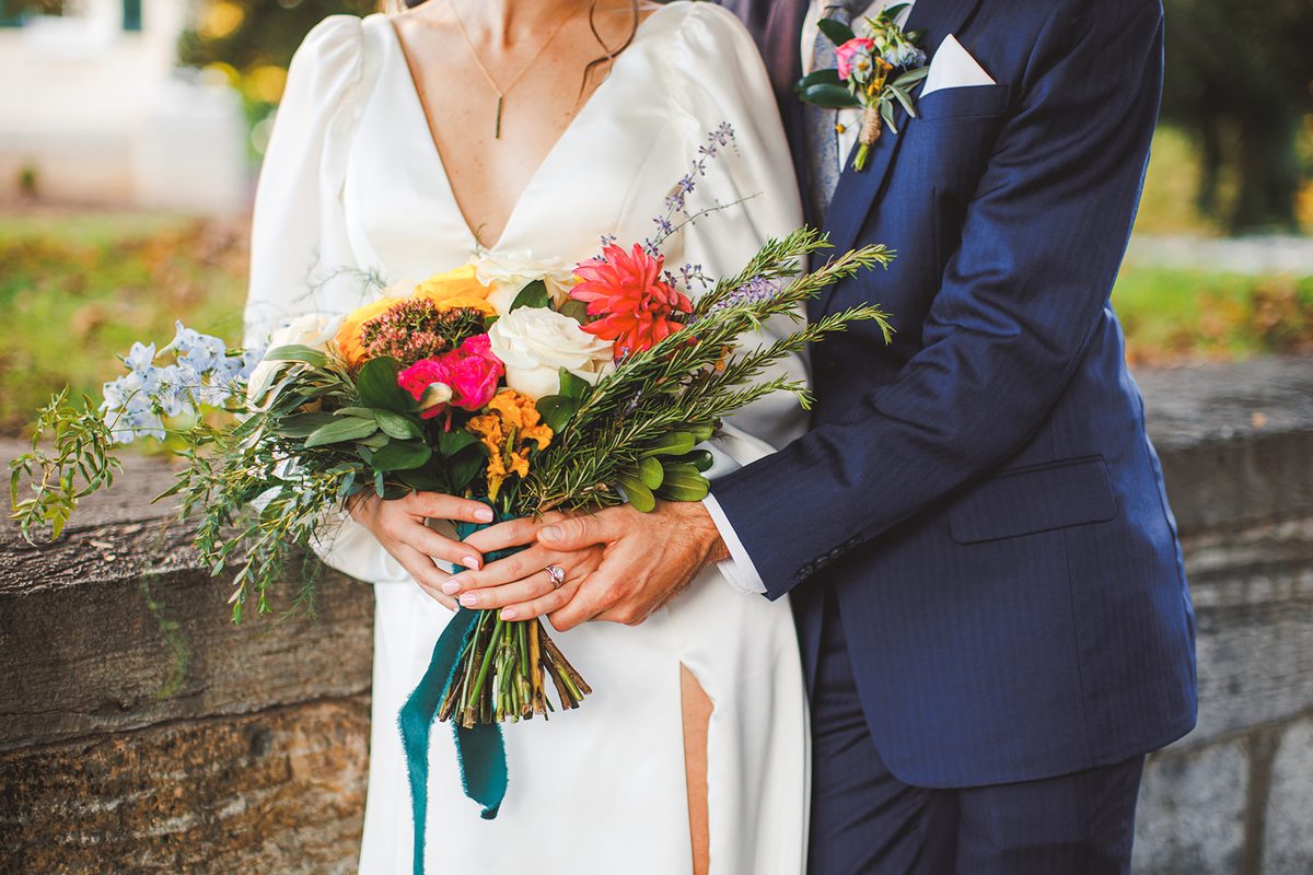 Close frame of Patrick's arms around Andrea from behind, both of their hands holding her bouquet of coral dahlias, cream roses, and rosemary