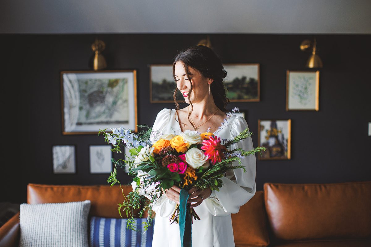 Andrea looking down at her bouquet of orange roses, pink dahlias, and pale blue delphinium, standing against the dark gallery wall of the lounge
