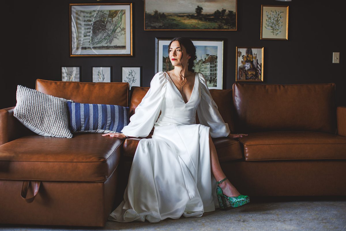 Andrea seated on the leather sofa beneath the framed gallery wall, her satin sleeves and skirt spread around her, one green platform heel showing