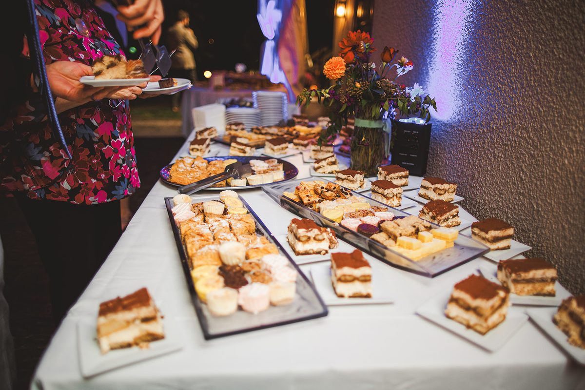 A long dessert table lined with tiramisu, pastries, and cookies under evening light, a guest filling a plate at the near end