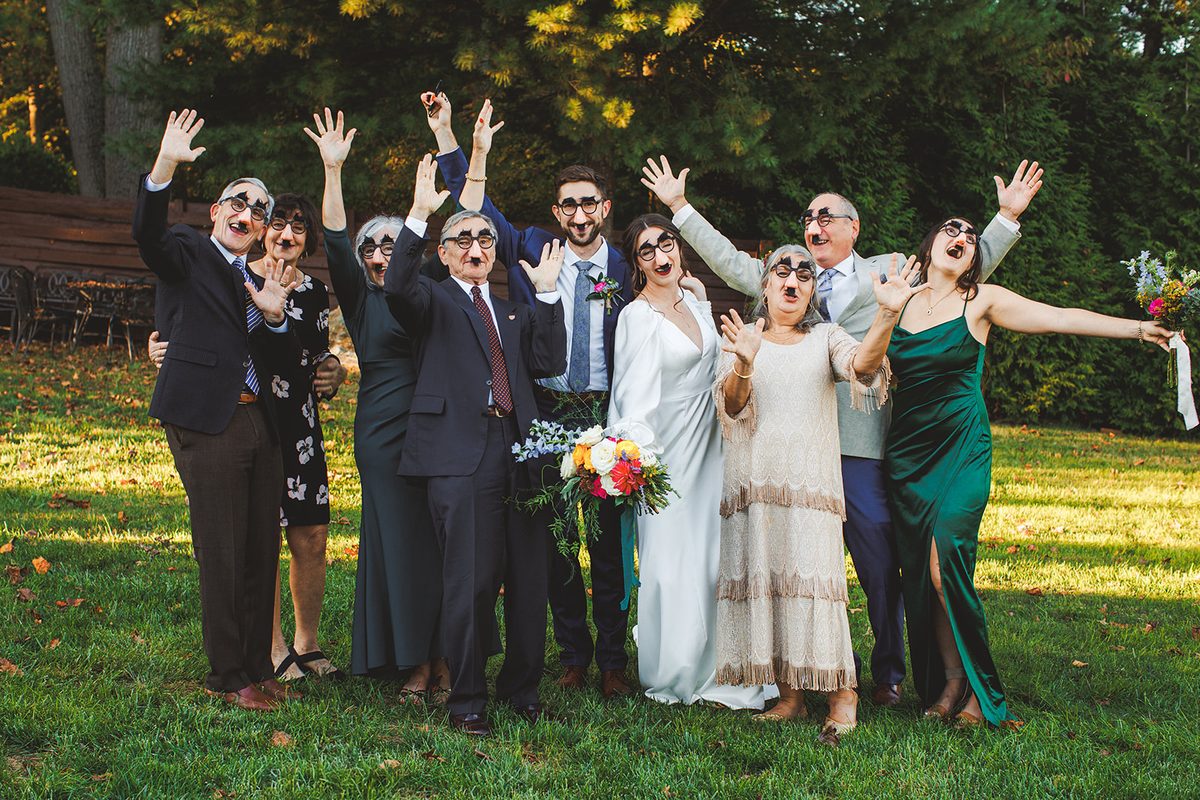 Andrea and Patrick surrounded by family on the lawn, every single person wearing Groucho glasses and mustaches with their hands thrown in the air, golden light behind them