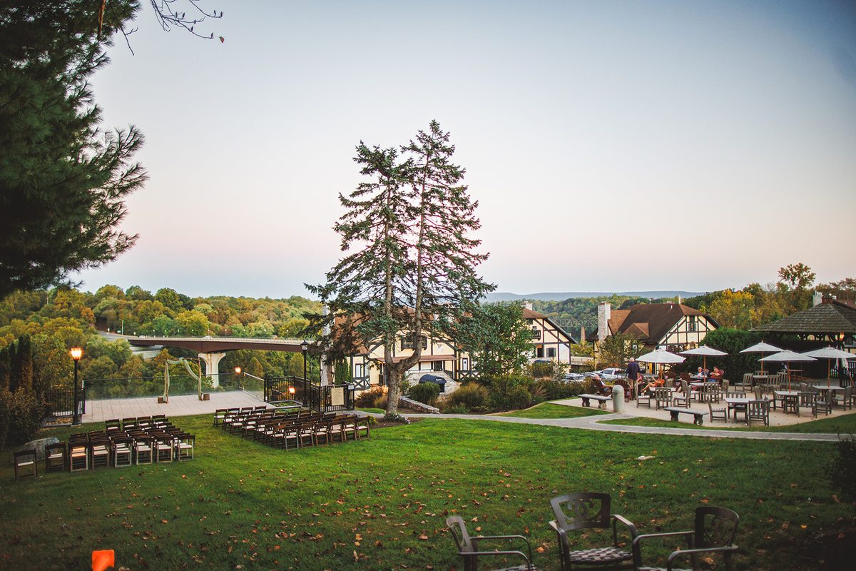Wide view of the Bavarian Inn grounds at dusk: the ceremony chairs and draped arch on the lawn, the half-timbered chalets and umbrella terrace to the right, and the bridge over the Potomac beyond