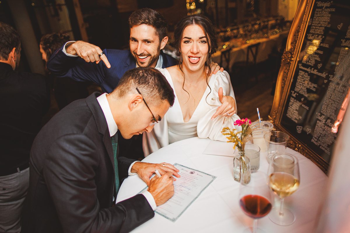 A witness signing the marriage license while Andrea and Patrick point at the paper and mug for the camera