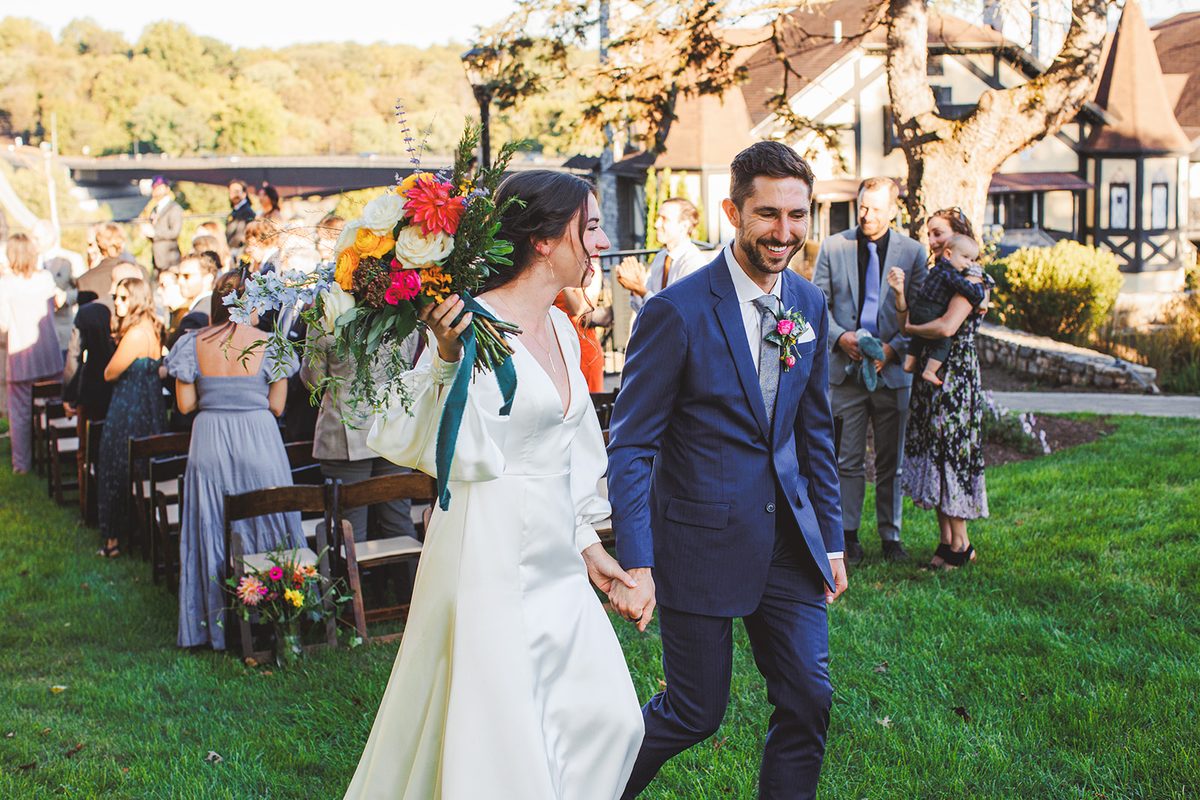 Andrea and Patrick walking back up the grass aisle hand in hand, both grinning, her bouquet raised past the applauding guests
