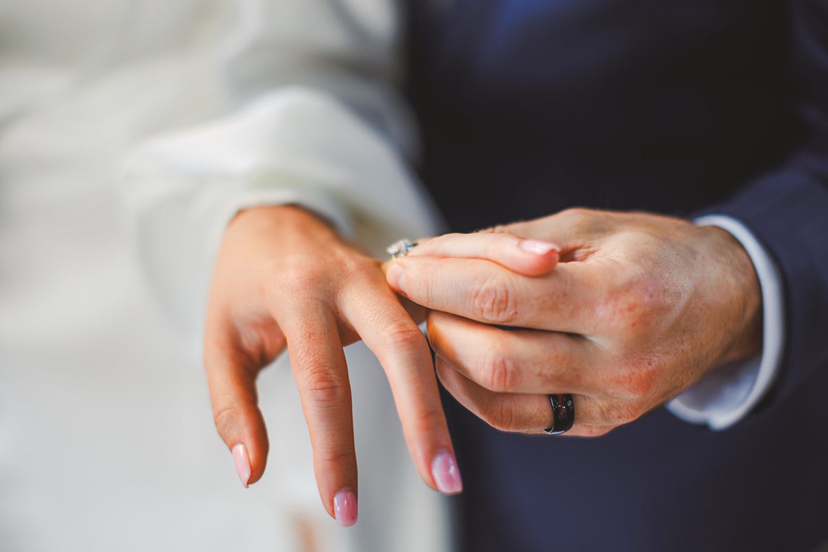 Patrick holding Andrea's hand in both of his, her engagement ring and his black band close together against her white sleeve