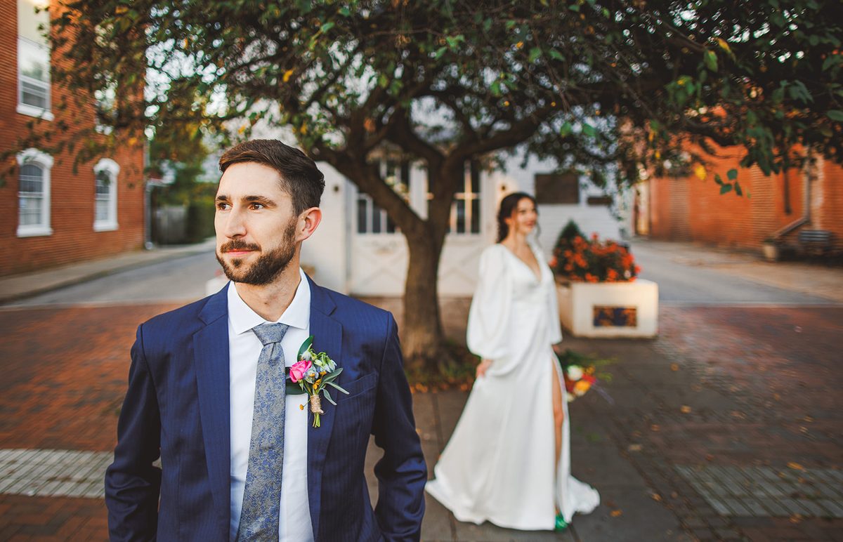 Patrick in the foreground in his navy suit and floral boutonniere looking off down the street while Andrea stands soft-focused by the tree behind him