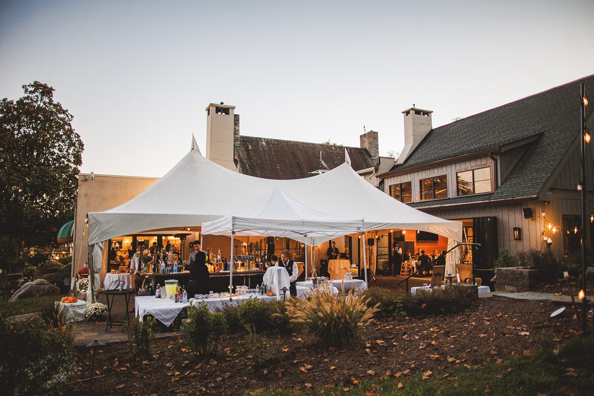 The white bar tent glowing warm at dusk beside the inn, bartenders working and guests gathered under strings of lights