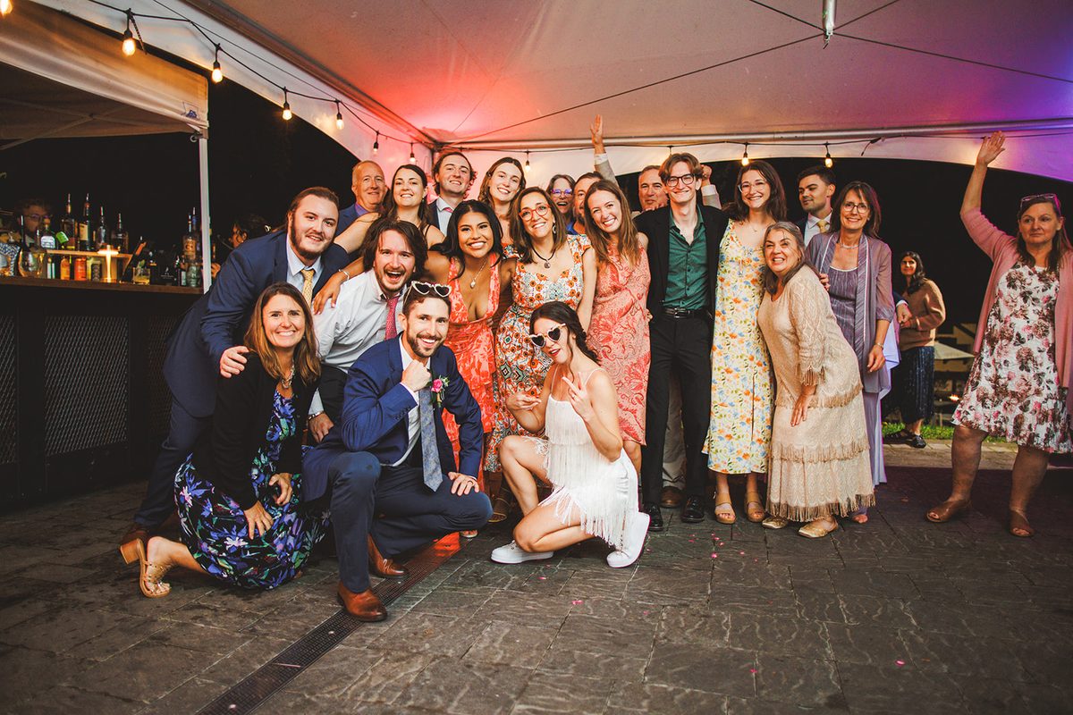 A crowd of guests packed together under the tent in pink and purple light, arms around each other, all shouting toward the camera