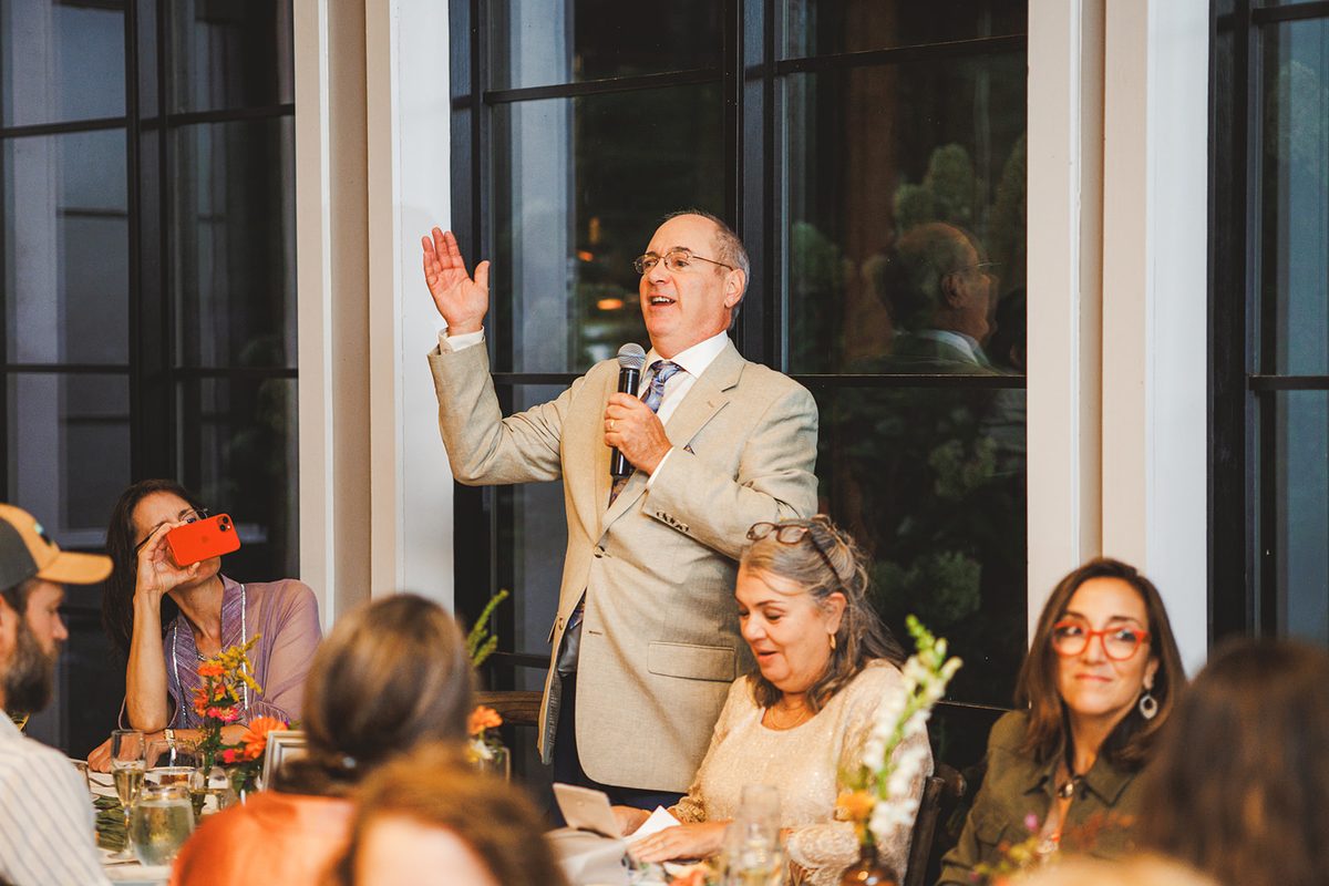 A toast being delivered by the tall windows of the brewhouse room, guests laughing along