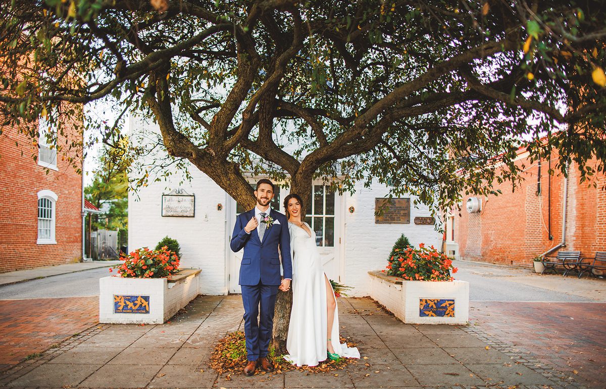 Andrea and Patrick standing together at the base of the great corner tree, its branches spreading overhead across the brick and white storefront