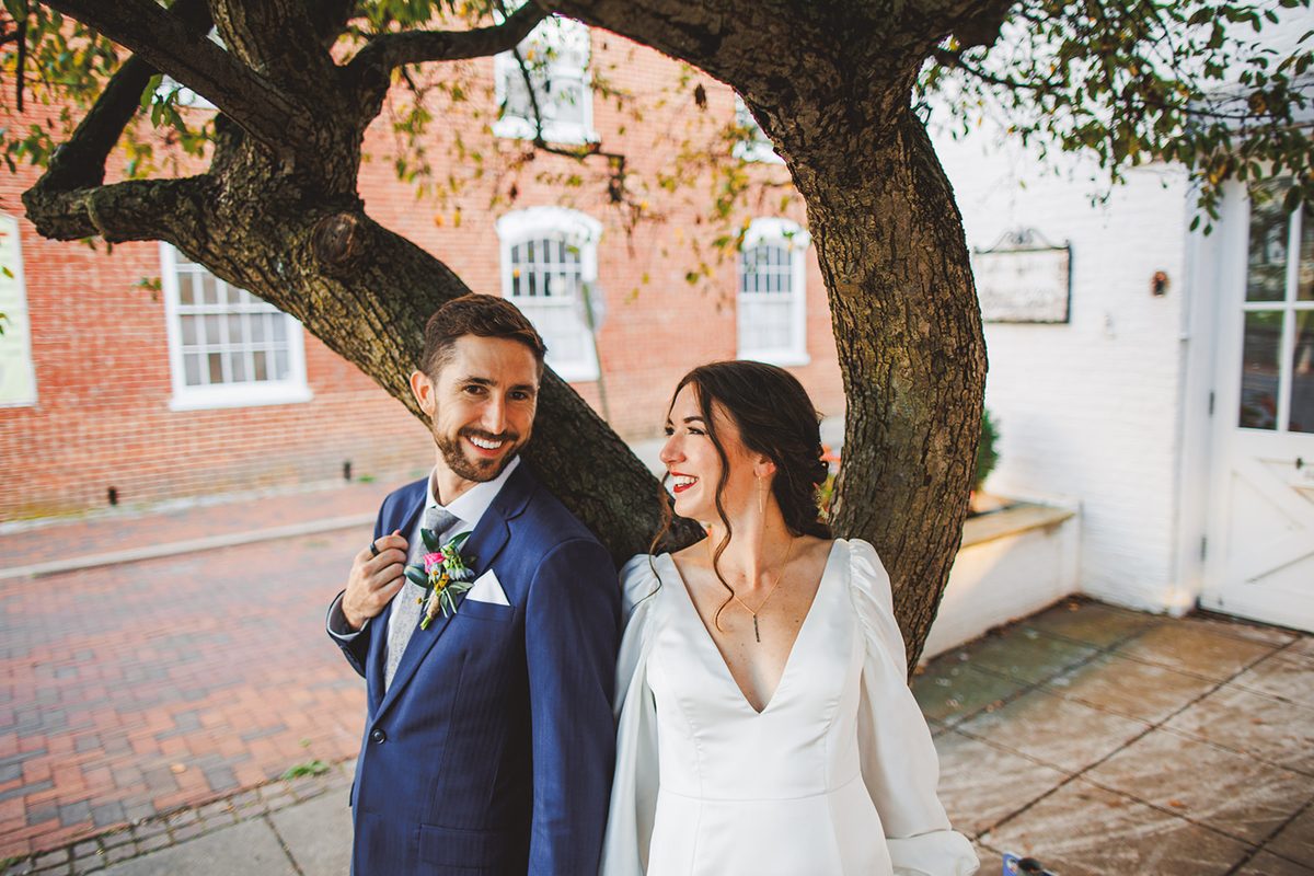 Andrea laughing up at Patrick beneath the low branches of an old tree, a red brick building behind them