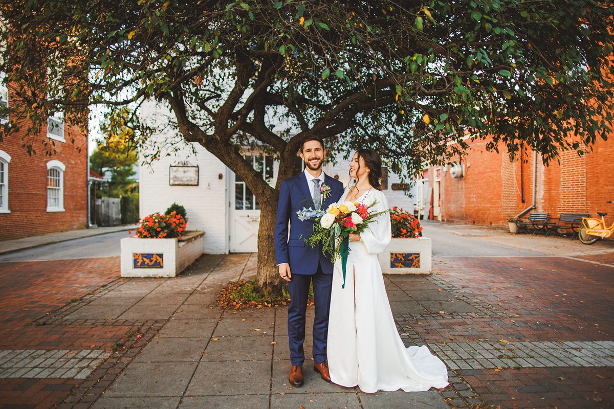 Andrea and Patrick standing hand in hand beneath the wide canopy of the old corner tree, brick street and planters of orange flowers on both sides
