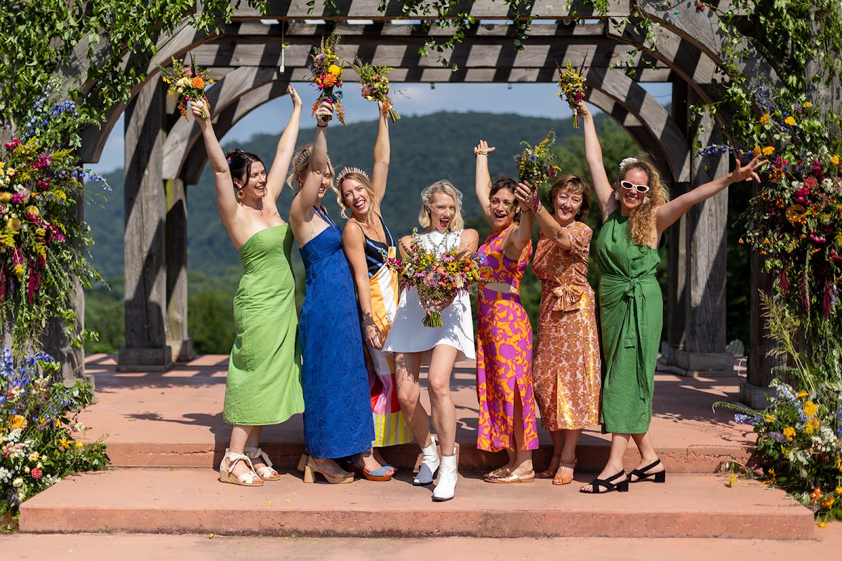 Annie and her bridal party in green, blue, orange, and pink dresses cheering under the pergola with bouquets raised