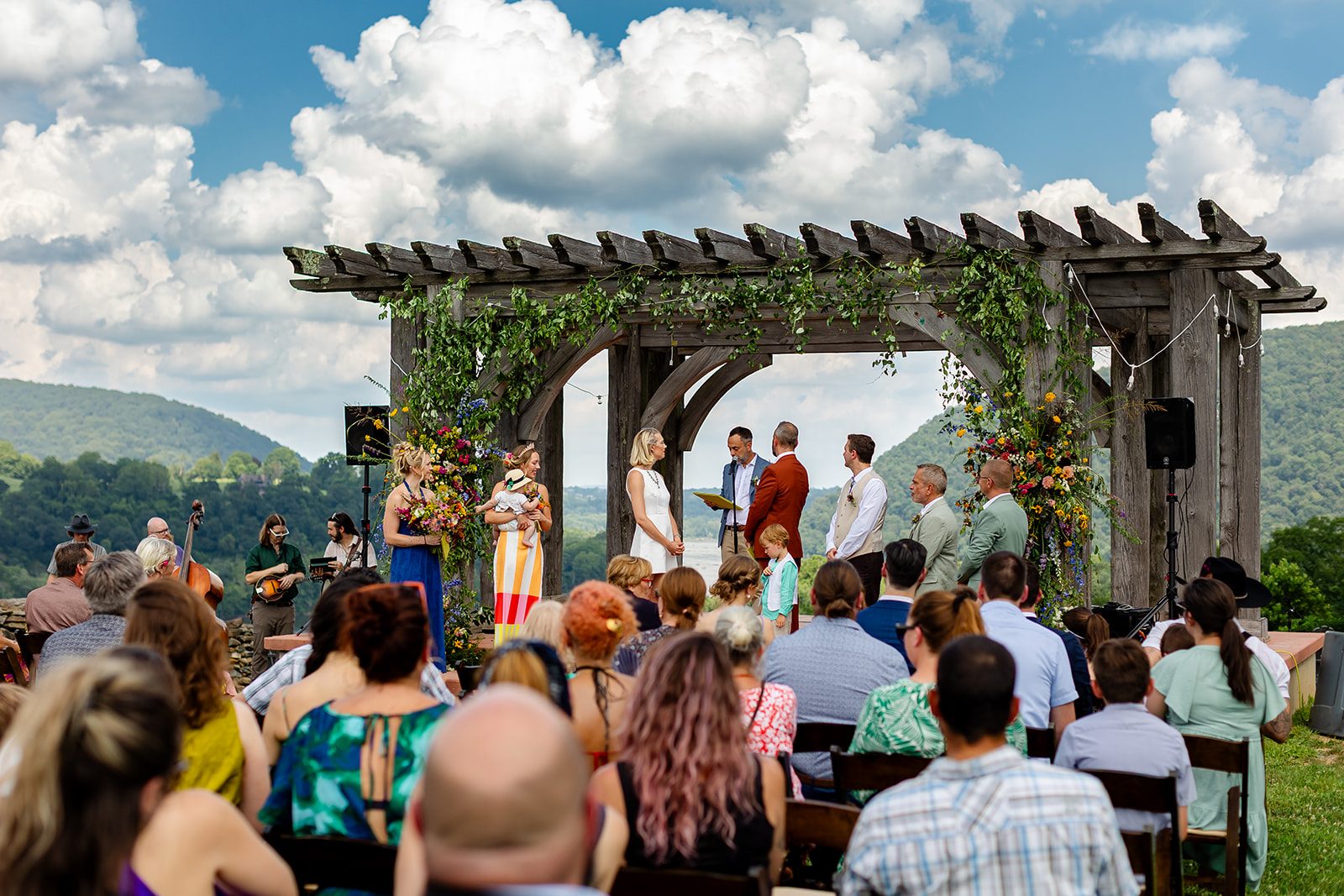 A ceremony under a broad timber pergola hung with greenery and wildflowers, seen from among the seated guests, the river water gap visible through the arch and a string band playing off to one side