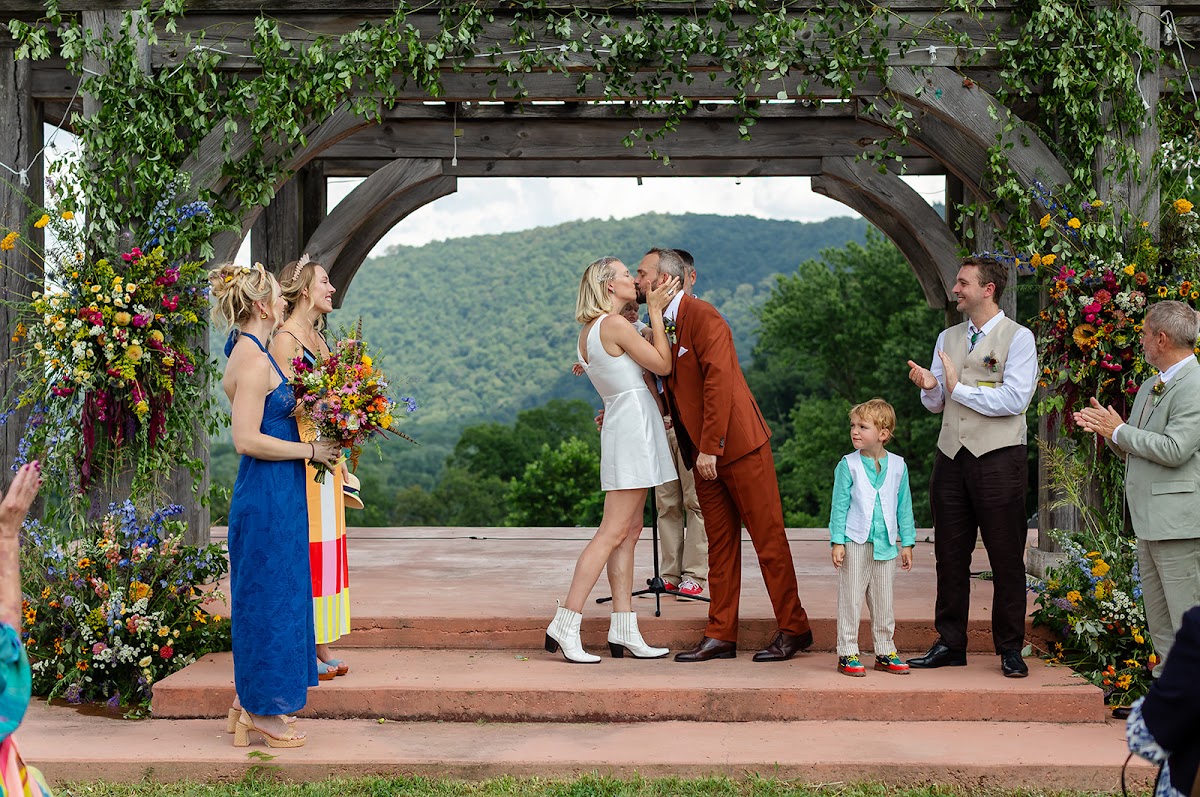 Annie and Thomas kissing at the top of the pergola steps as the wedding party and guests applaud, wildflower arrangements on both sides