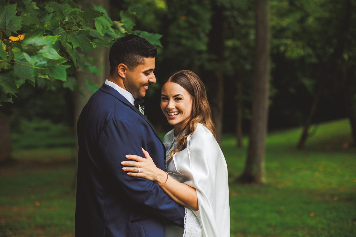 Bailey laughing at the camera from inside Youssef's arms beneath the tall trees, her satin cape catching the light