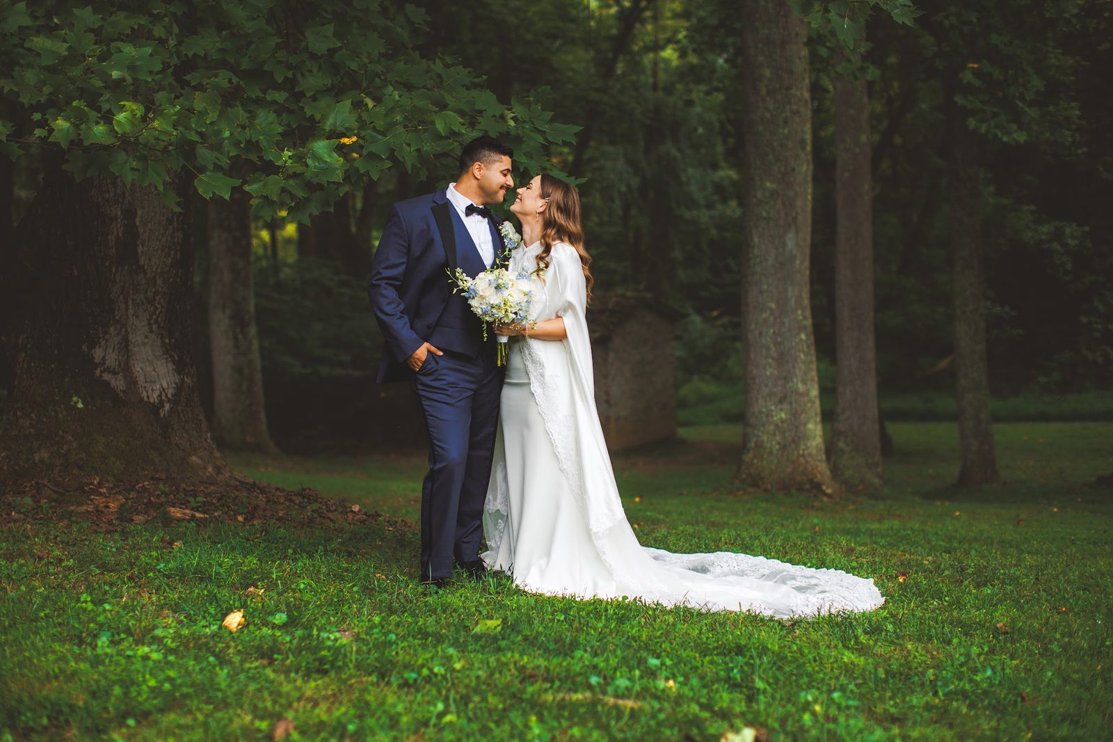 Bailey and Youssef nose to nose beneath towering trees at a Brinklow, Maryland estate, her white satin cape and lace-edged train spread across the grass