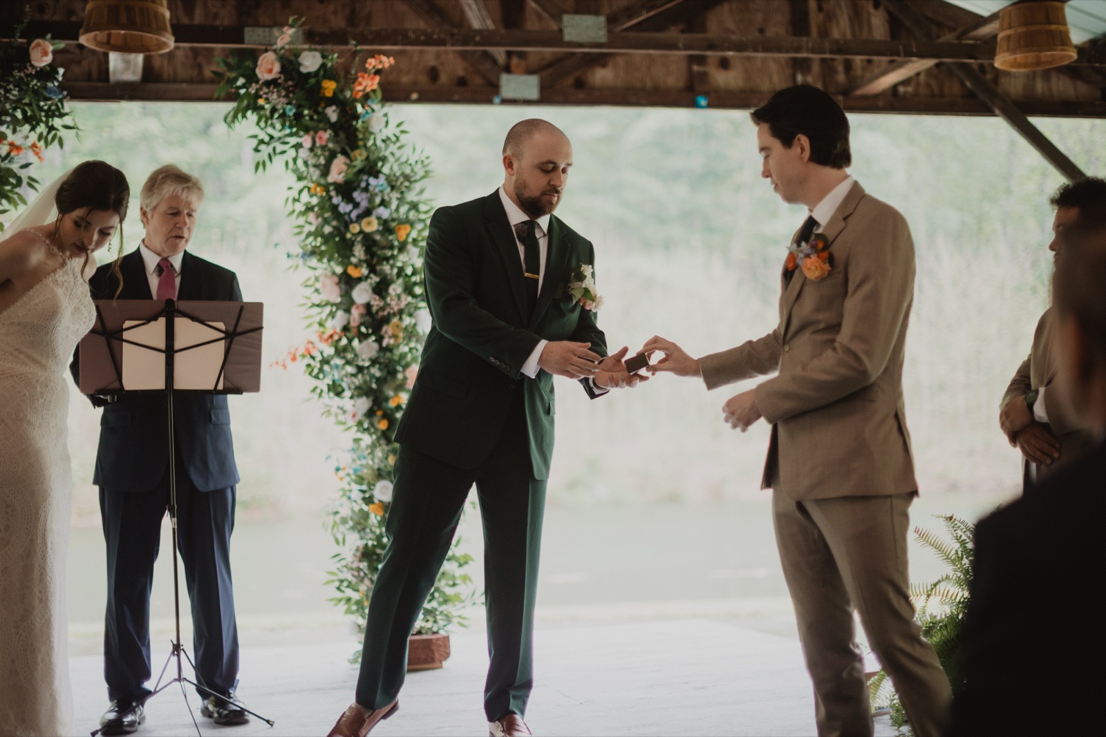 A ring being slid onto a finger under a floral arch, both of them watching their own hands, the officiant holding an open book just behind