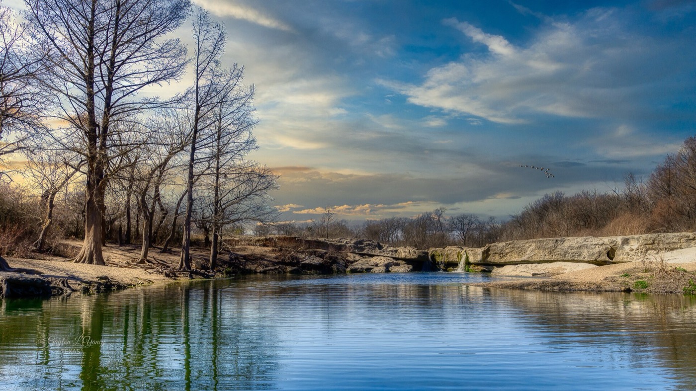 A calm river reflecting a dramatic banded sky at dusk, bare cypress trees along the near bank and a low bridge crossing in the distance