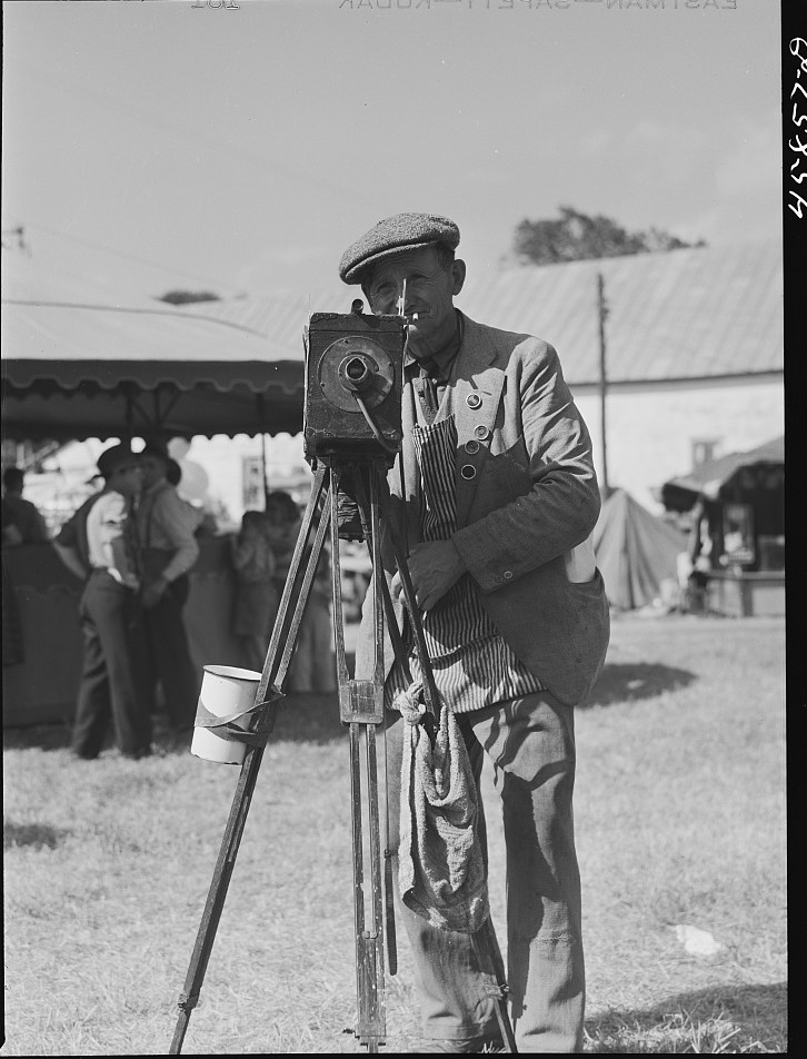 A tintype photographer in a flat cap standing beside his wooden plate camera on a tripod at a Vermont fair in 1941, a developing bucket hanging from the tripod legs