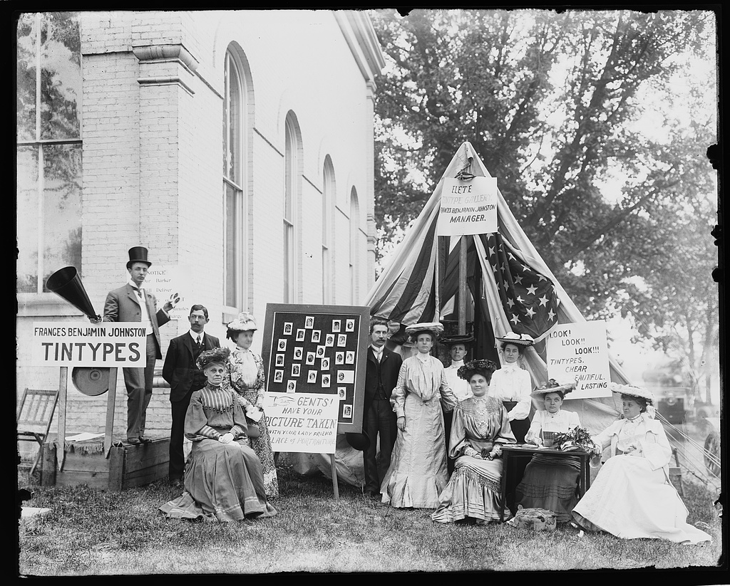 A photographer's tent at a county fair in 1903, hung with flags, beside a large board of sample tintype portraits and hand-lettered signs reading Frances Benjamin Johnston Tintypes