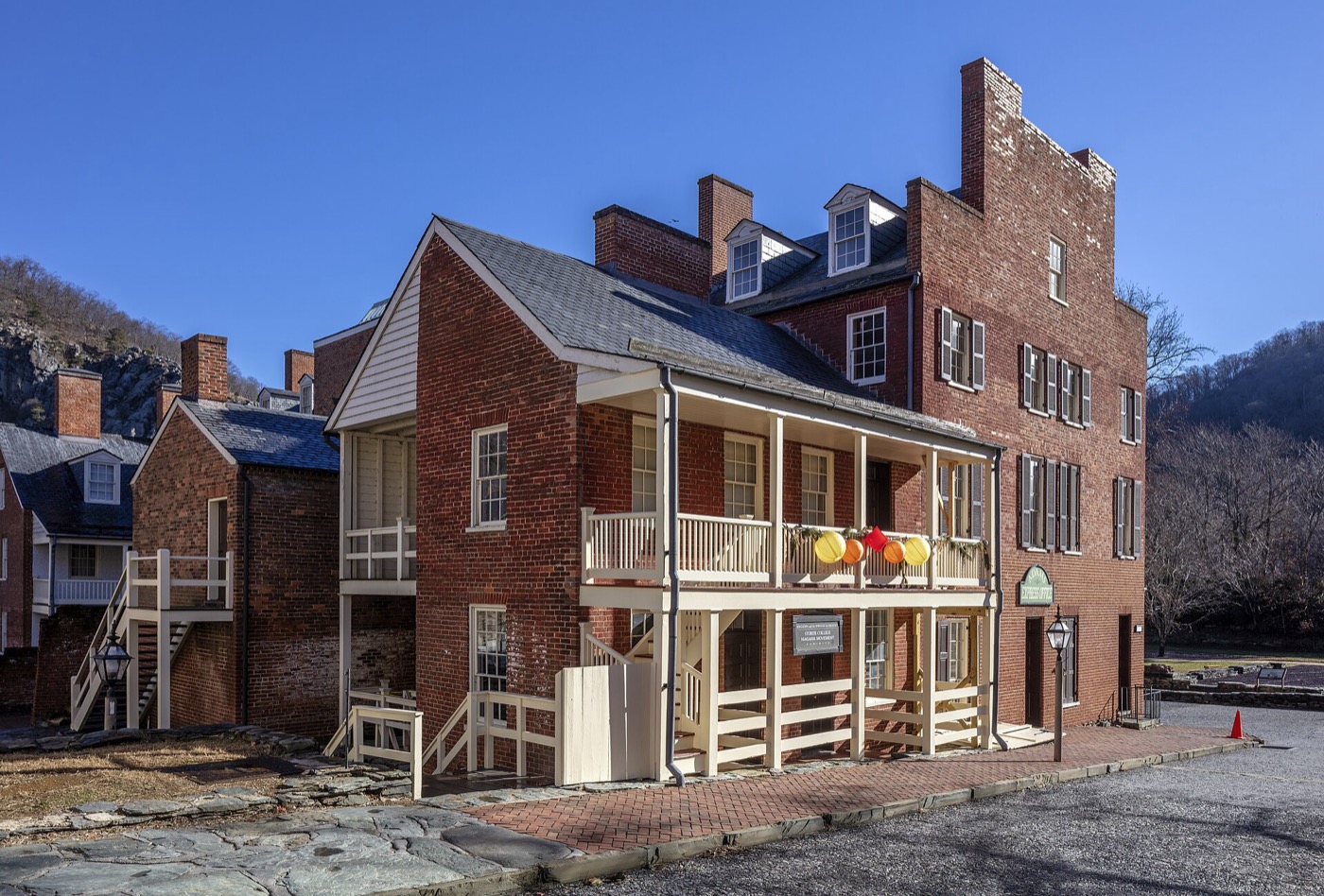 Brick and stone buildings stepping up Lower High Street in Harpers Ferry under a clear sky, wooded hillside rising behind the rooftops