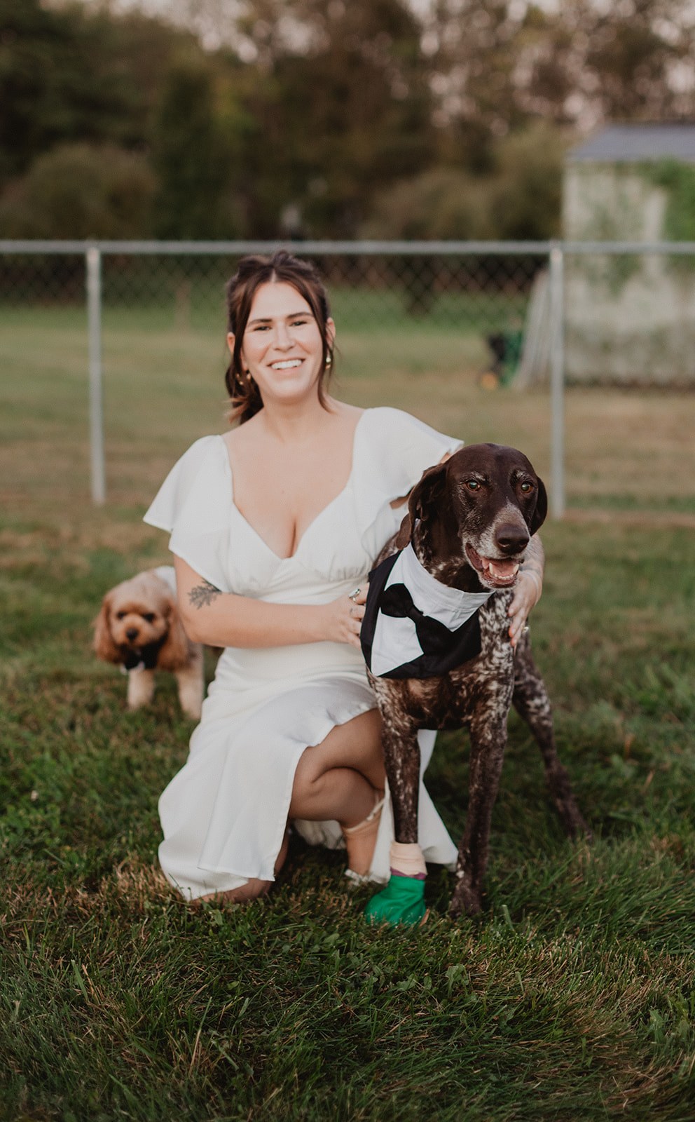 Michaela crouched in the grass in her white dress with one arm around the pointer in his tuxedo bib, the small tan doodle in a bow tie standing at her other side