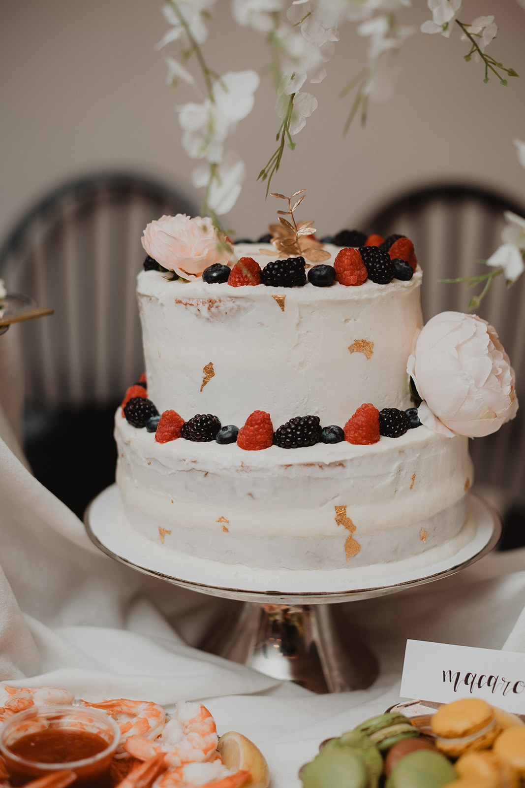A two tier semi naked wedding cake ringed with blackberries, raspberries and strawberries on a silver stand, with macarons behind it