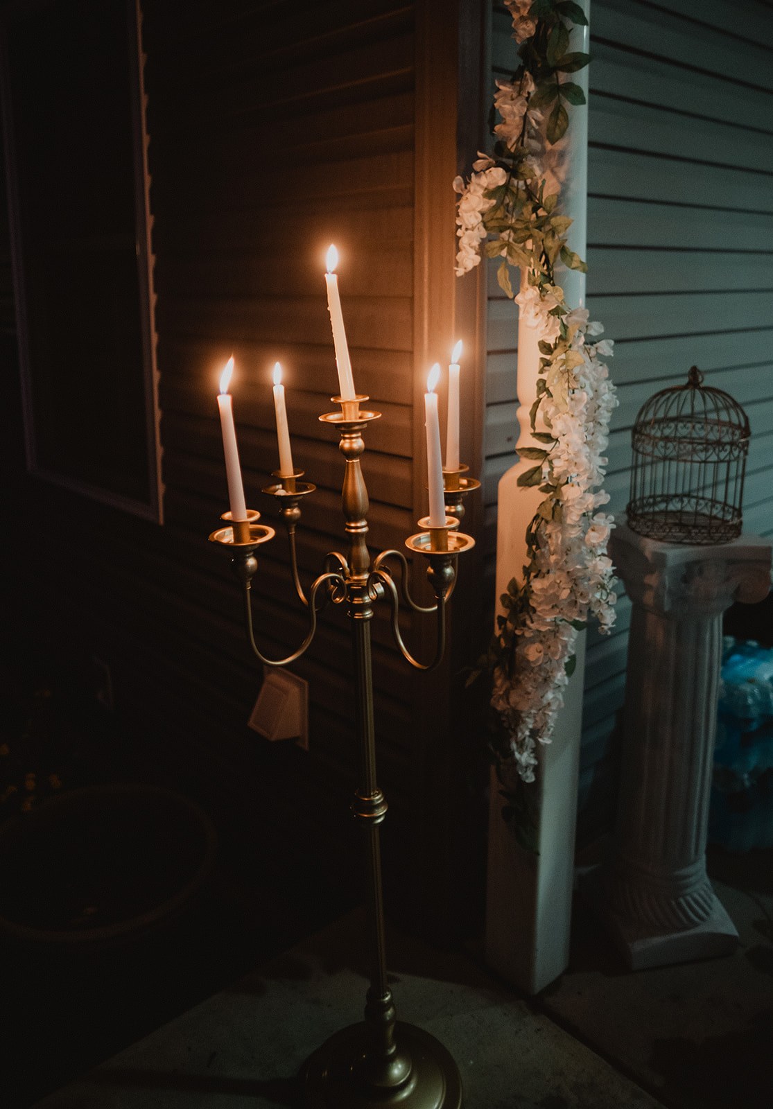 A brass candelabra of burning tapers on a dark patio at night, white orchids and a birdcage behind it