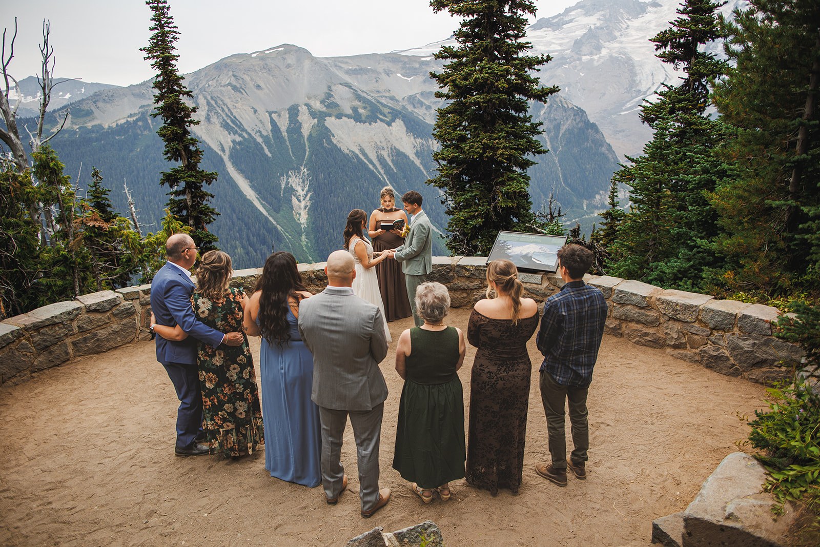 The ceremony at Emmons Vista seen from behind the guests: eight people standing in a half circle on a stone terrace facing Michaela, AJ, and the officiant, with Mount Rainier's glaciers filling the background