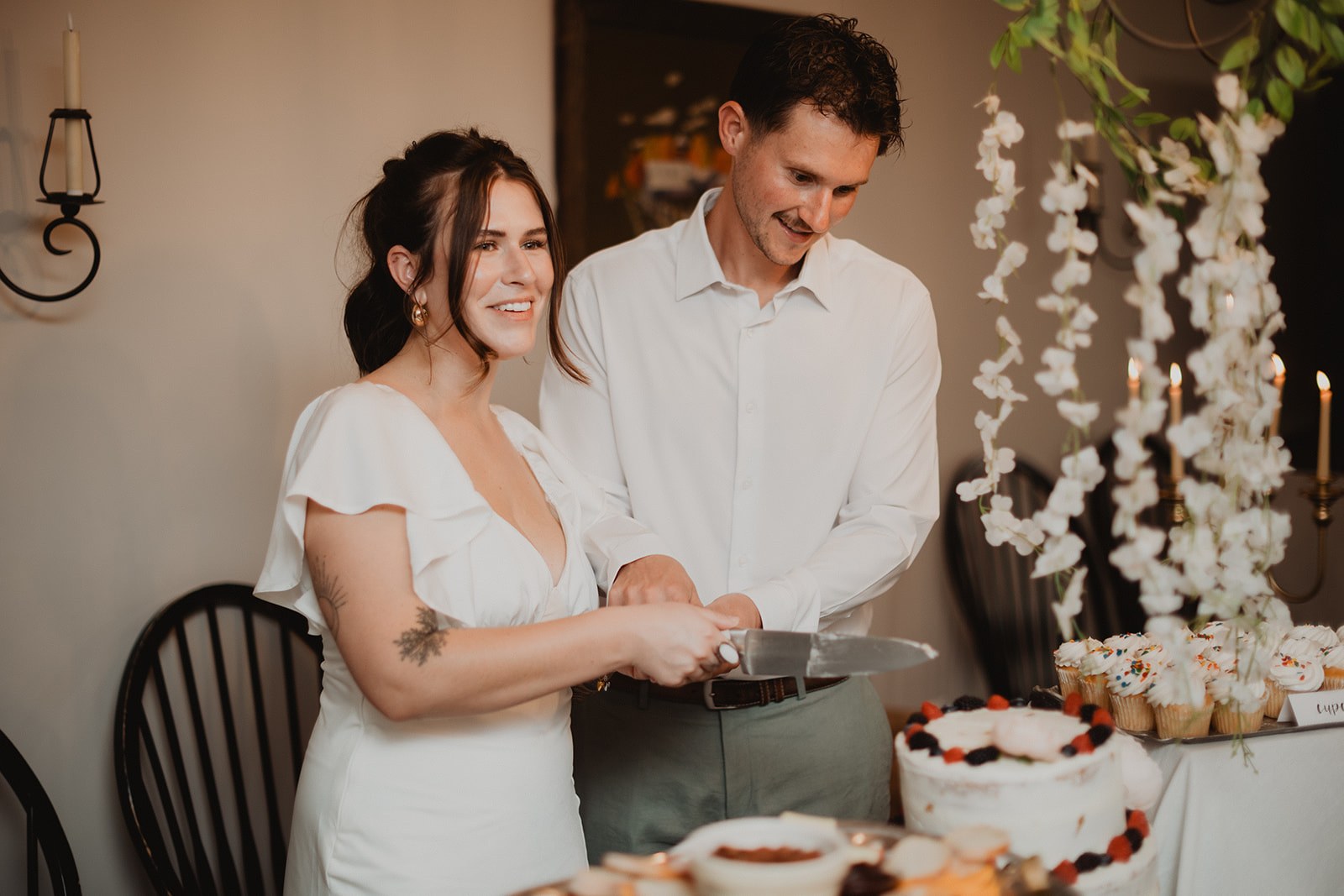 Michaela and AJ cutting into the cake together indoors, long strands of white orchids hanging on the wall behind them