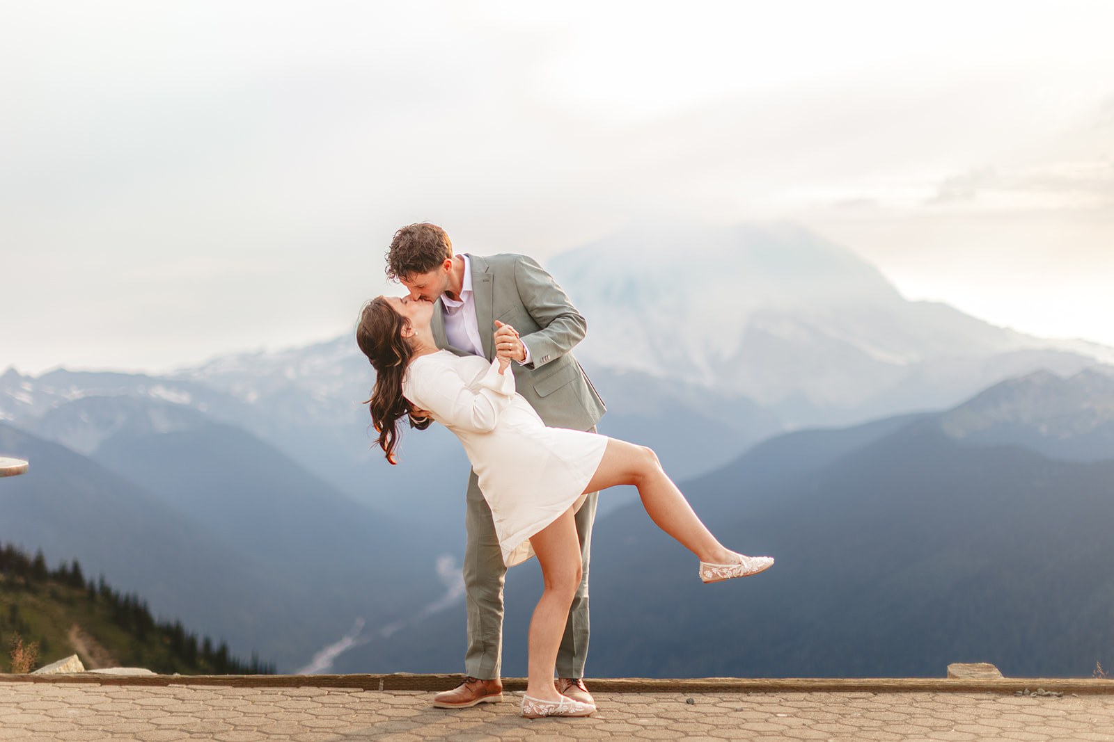 AJ dipping Michaela into a kiss on the summit paving at sunset, one of her feet kicked up behind her, Mount Rainier hazy and enormous across the valley