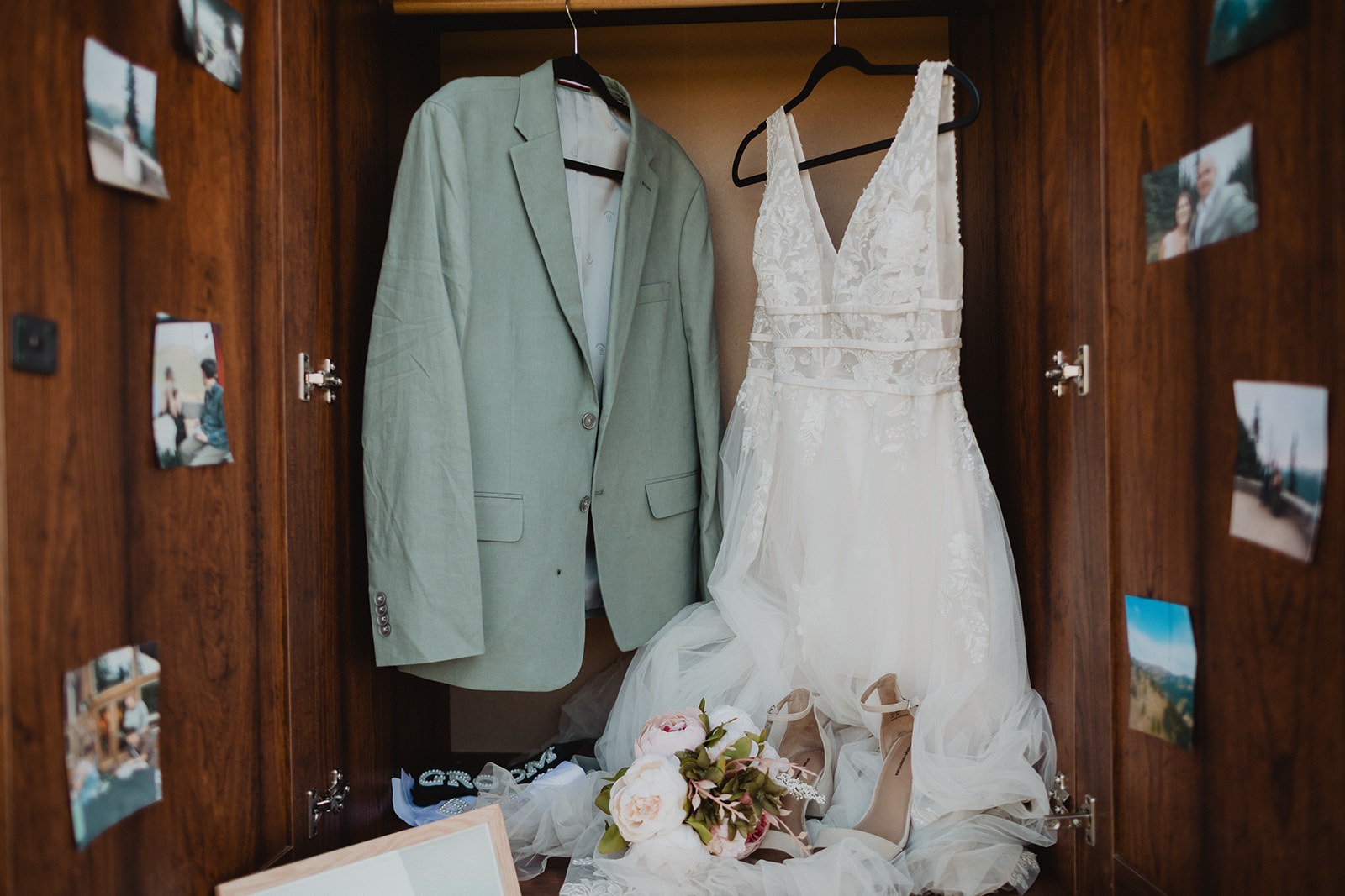 Michaela's wedding dress and AJ's sage linen jacket hanging inside an open wooden cabinet, with printed photographs from their Mount Rainier elopement pinned all around the doors