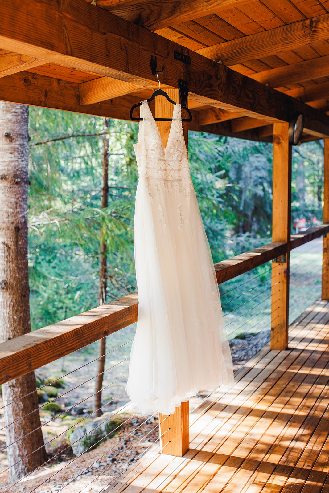 Michaela's lace and tulle wedding dress hanging from a beam on the cabin porch, sunlight across the deck boards and evergreens behind the rail