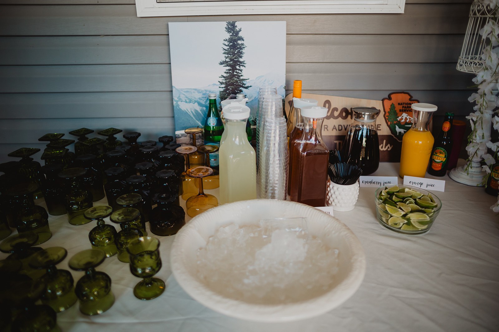 A drinks table on the patio with rows of green pressed glass tumblers, jugs of lemonade and iced tea, and a bowl of ice