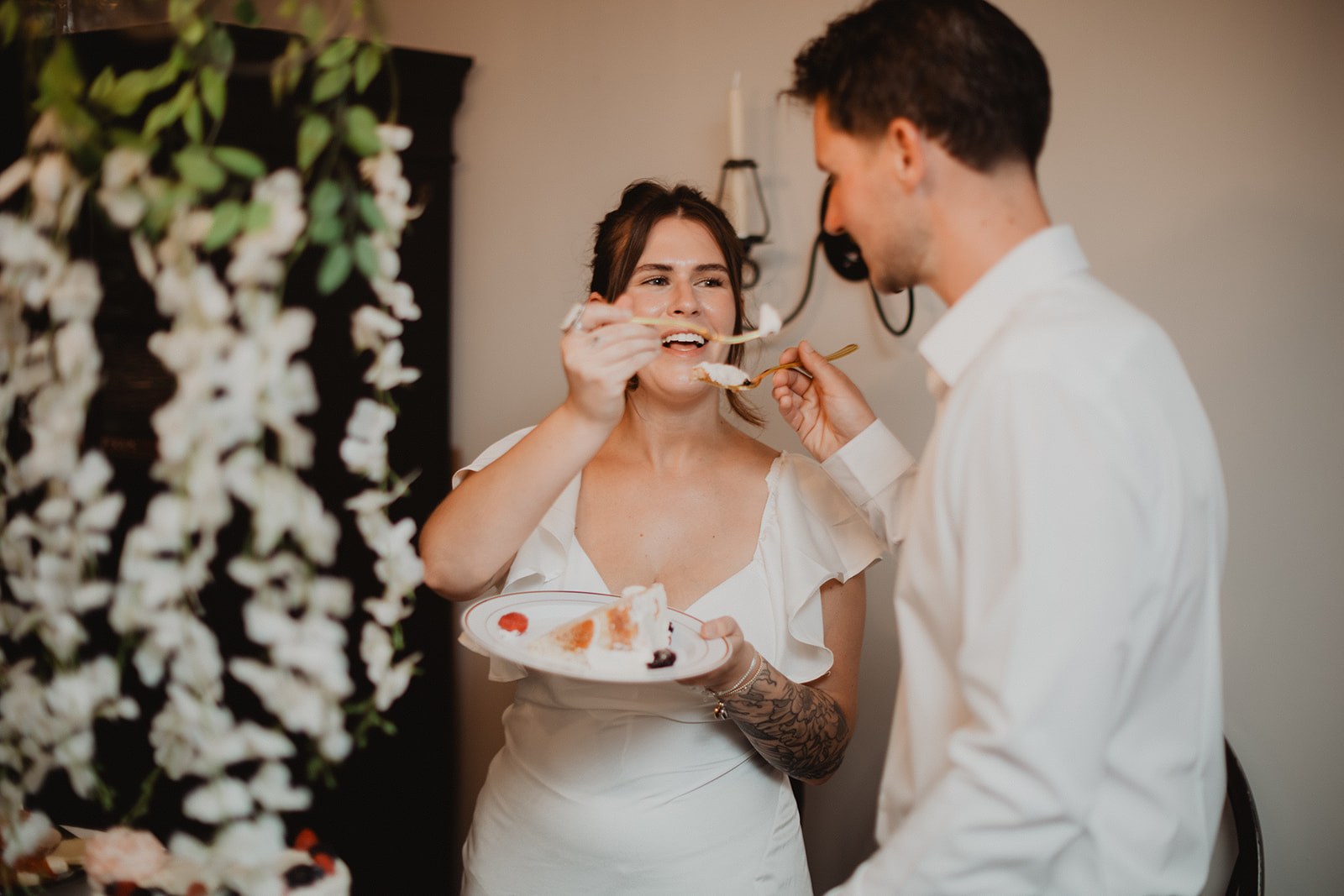 Michaela and AJ feeding each other the first bite of cake indoors, hanging strands of white orchids behind them and a brass wall sconce beside