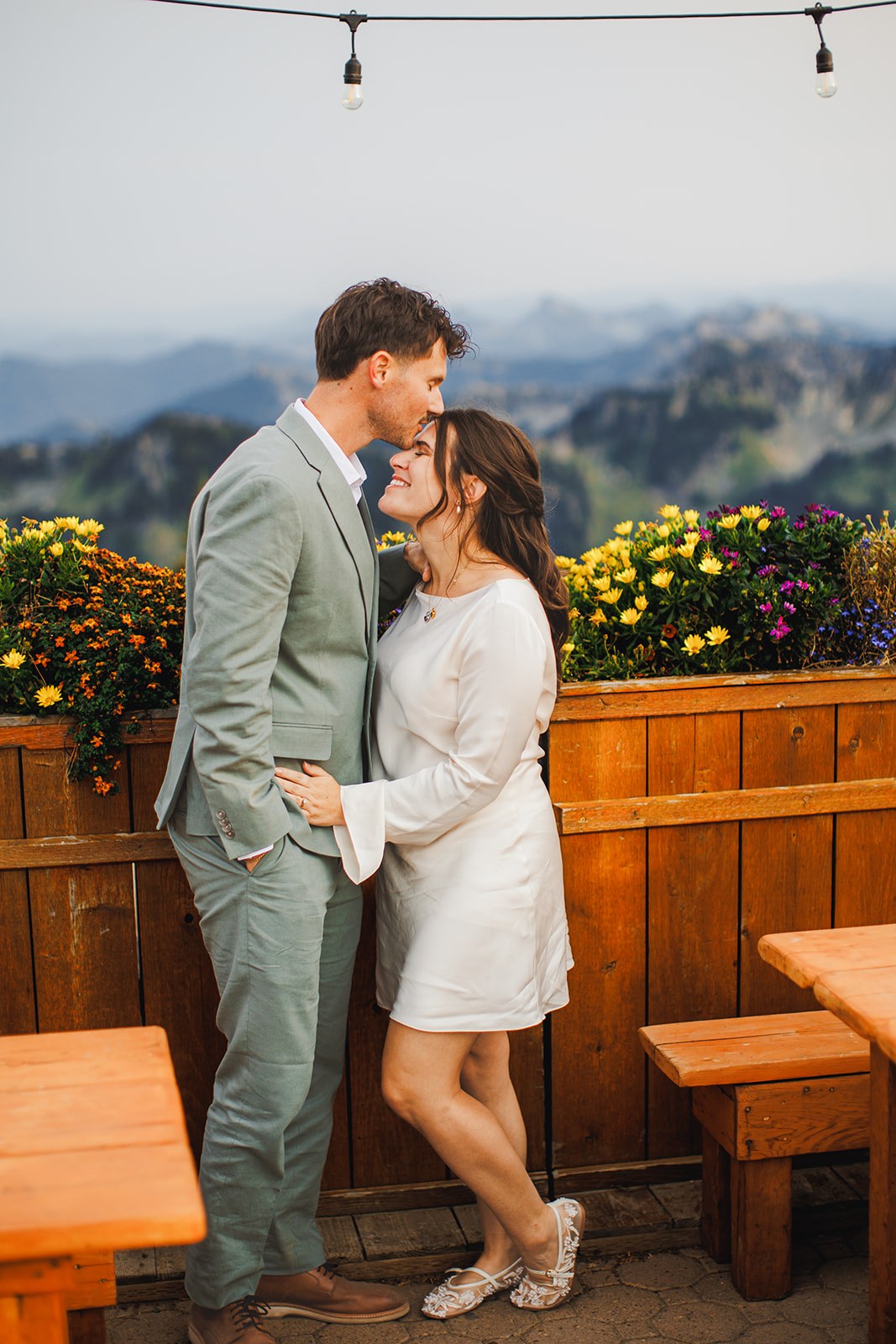 AJ kissing Michaela's forehead on the summit deck beside cedar planters full of marigolds and pansies, string lights overhead