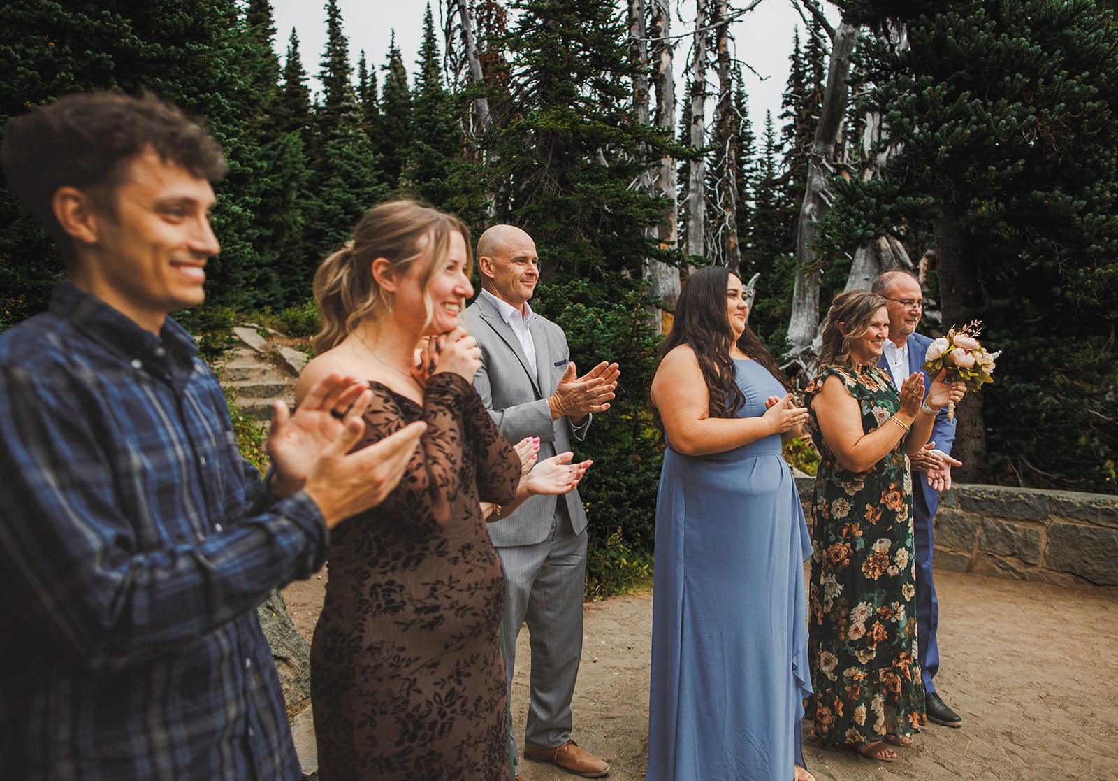 The line of guests applauding at the end of the ceremony, six of them shoulder to shoulder against the firs, one still holding the bouquet