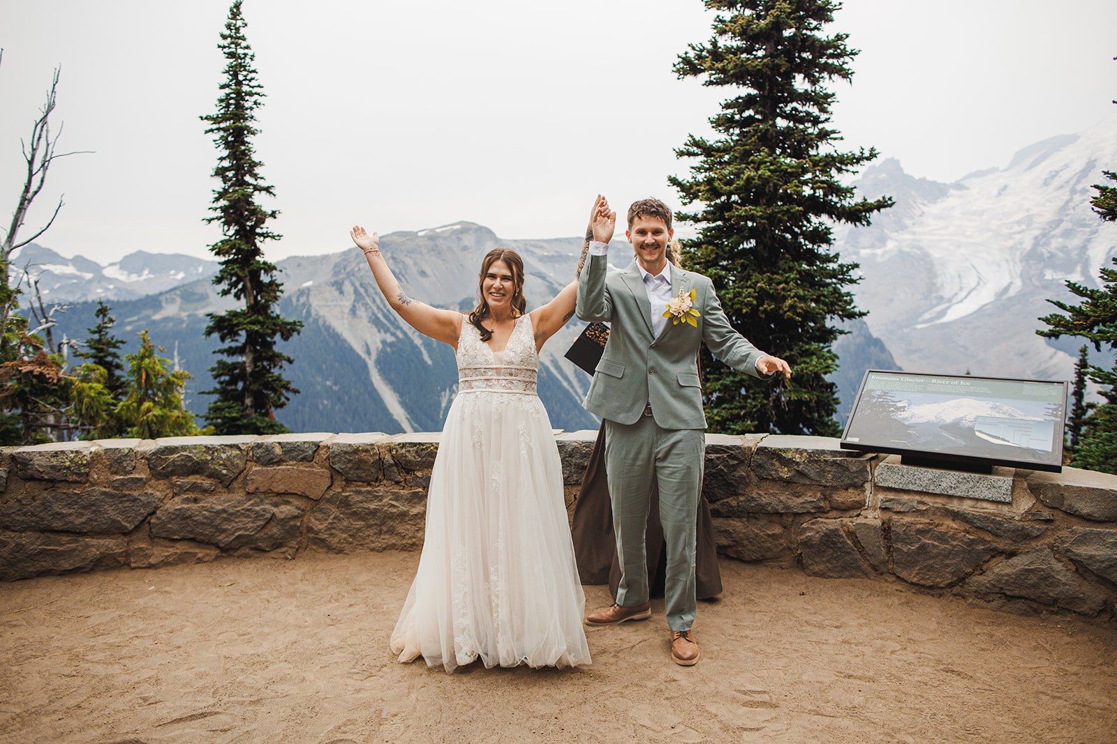 Michaela and AJ throwing both hands in the air the moment they are married, alone on the stone terrace with the glaciated ridges of Mount Rainier behind them